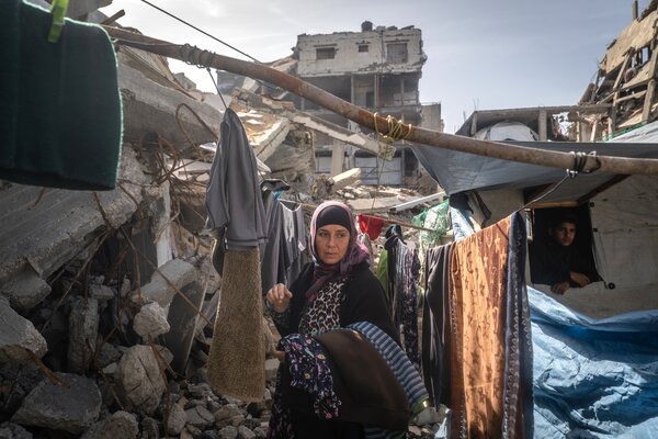 Woman and child in a makeshift shelter amid rubble, with hanging laundry and a heavily damaged building in the background.